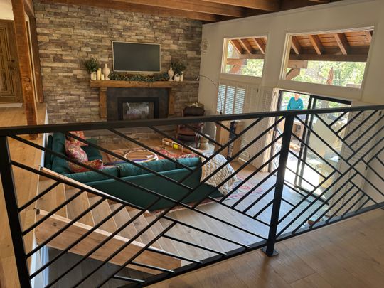 Interior view of a modern home showing a black metal stair railing overlooking a living room with stone fireplace, green sectional sofa, exposed wooden ceiling beams, and large windows