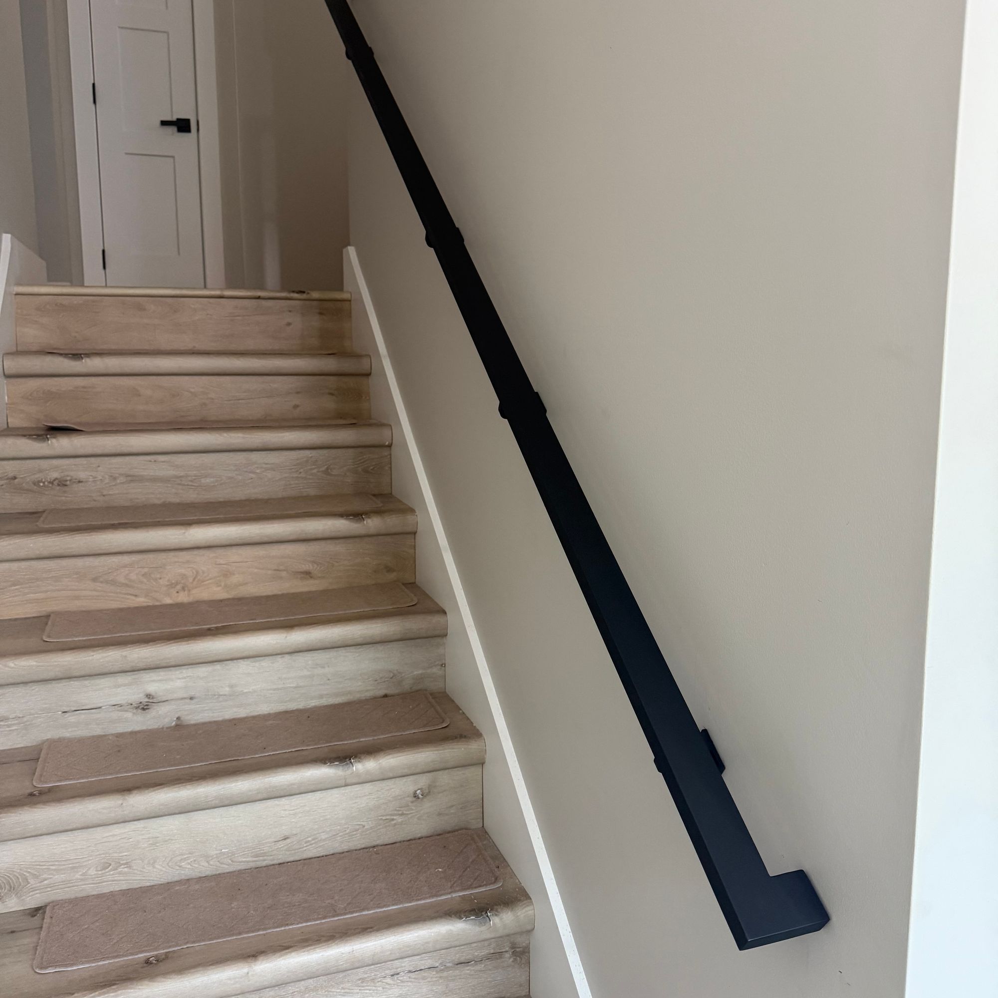 A staircase with light wood steps and white walls, featuring a black handrail mounted on the wall, leading up to a white door at the top of the stairs.