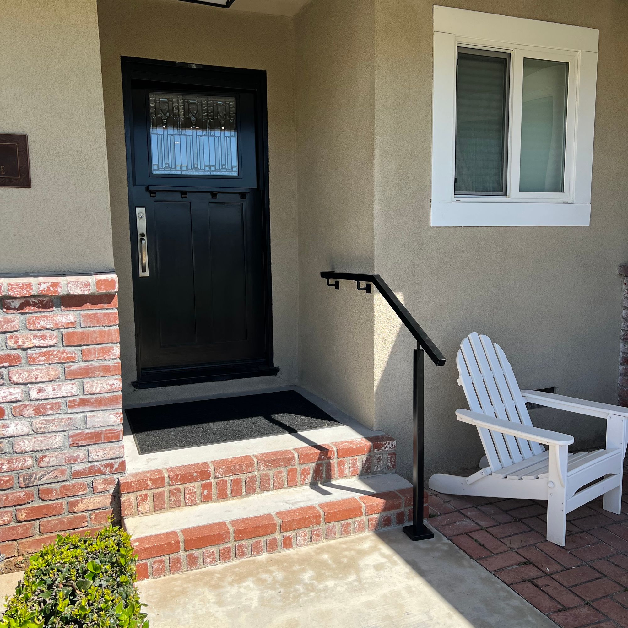 Front entrance of a house with black door, brick steps, white handrail, and white Adirondack chair on brick patio