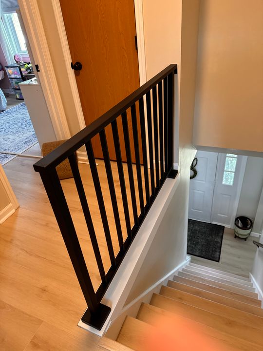 Interior staircase with black metal railing and vertical spindles, wooden steps, and white walls leading to upper floor of home