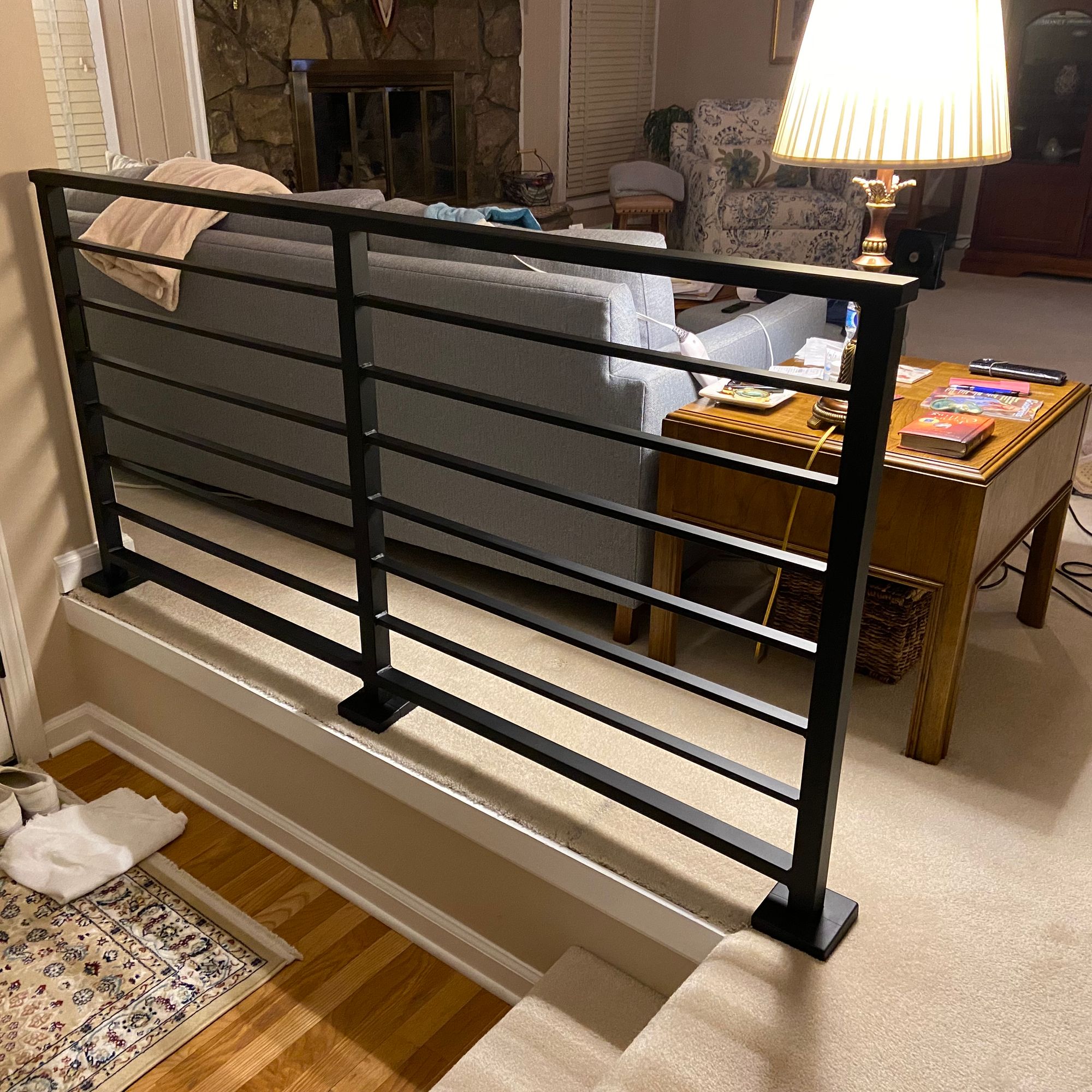 Interior view showing black metal stair railing overlooking a living room with beige carpet, wooden coffee table, gray sofa, stone fireplace, and television