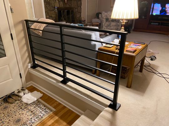 Interior view showing black metal stair railing overlooking a living room with beige carpet, wooden coffee table, gray sofa, stone fireplace, and television