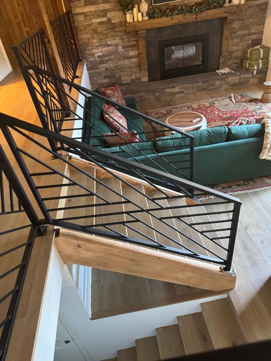 Modern staircase with black metal geometric railing overlooking a living room with stone fireplace and teal sectional sofa