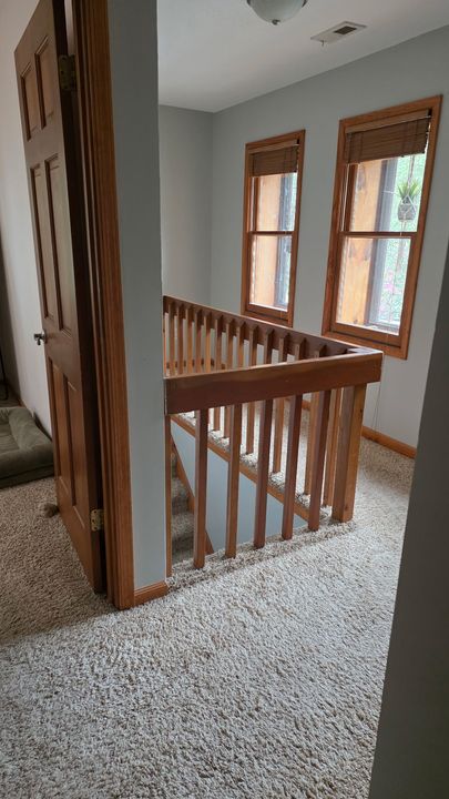 Interior hallway with wooden staircase railing, two windows with wood trim, beige carpet, and gray walls