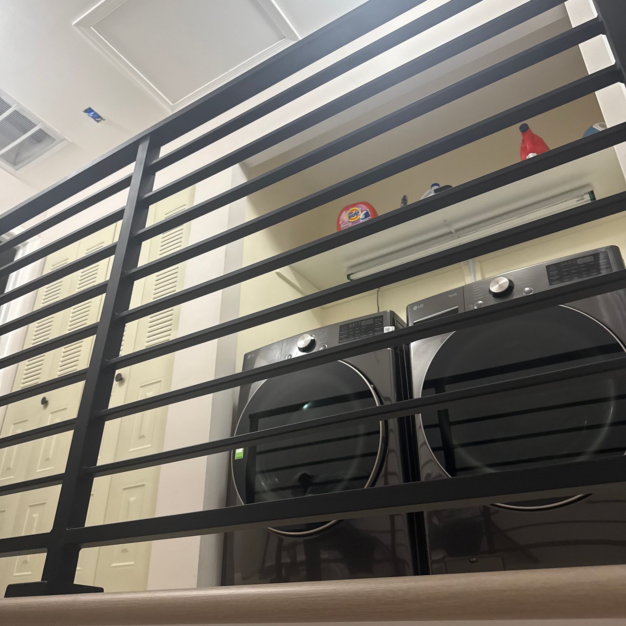 A loft bed with black metal railings positioned above a washer and dryer in a white room with built-in storage cabinets