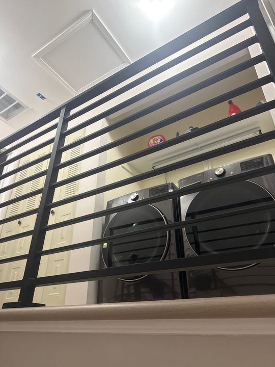 A loft bed with black metal railings positioned above a washer and dryer in a white room with built-in storage cabinets