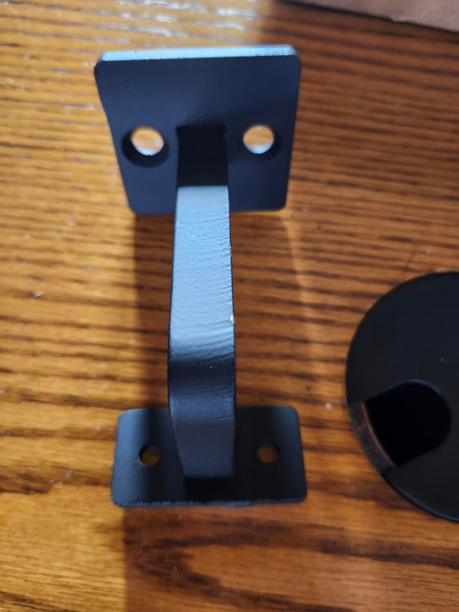 Black metal bracket or mounting hardware with two square plates connected by a curved metal strip, sitting on a wooden surface