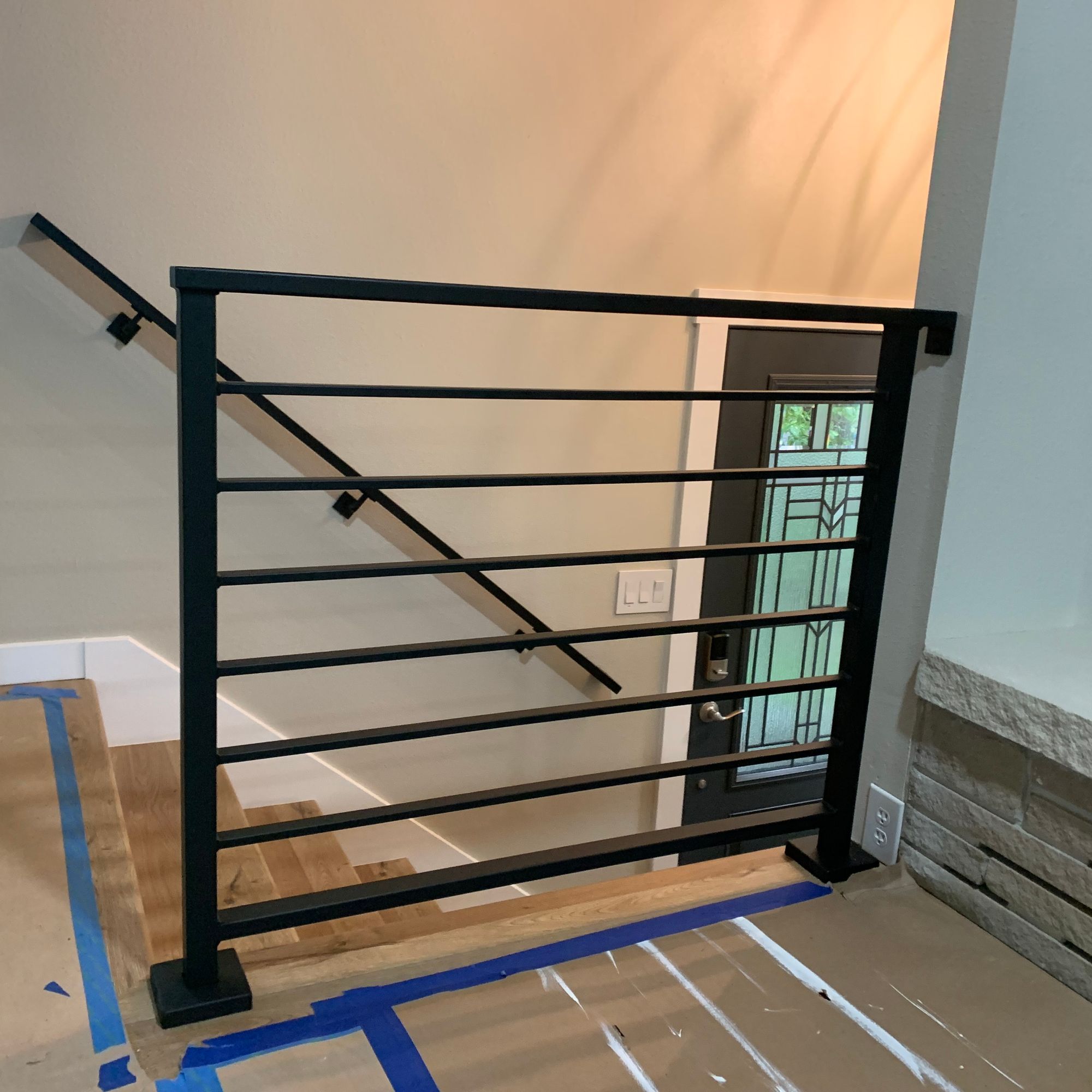 Modern black metal stair railing with horizontal bars installed in a home under construction, with protective blue tape on wooden floors and construction materials visible
