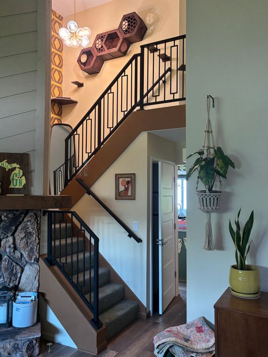 Interior view of a modern home showing a wooden staircase with black metal railings, decorative hexagonal wall art, hanging plants, and a stone fireplace