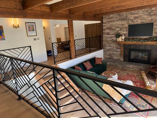 Interior view of a rustic modern home showing an upper level with exposed wooden beams, black metal railings with geometric patterns, a green sectional sofa below, stone fireplace with mounted TV, and warm lighting throughout the open-concept space.