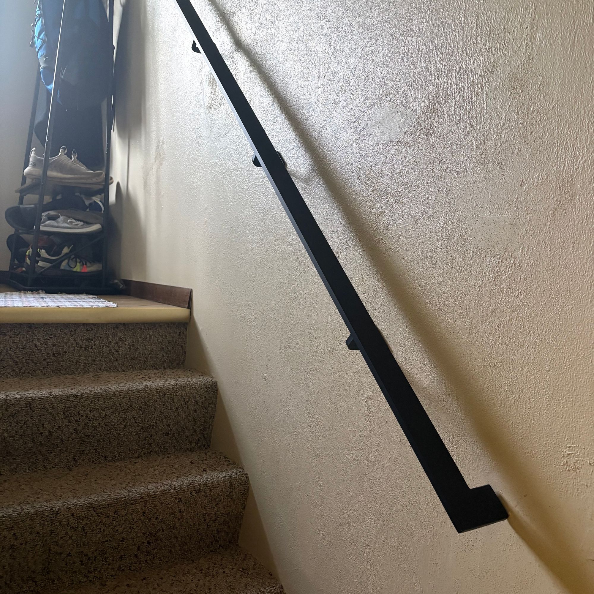 A black metal handrail mounted on a white wall alongside carpeted stairs