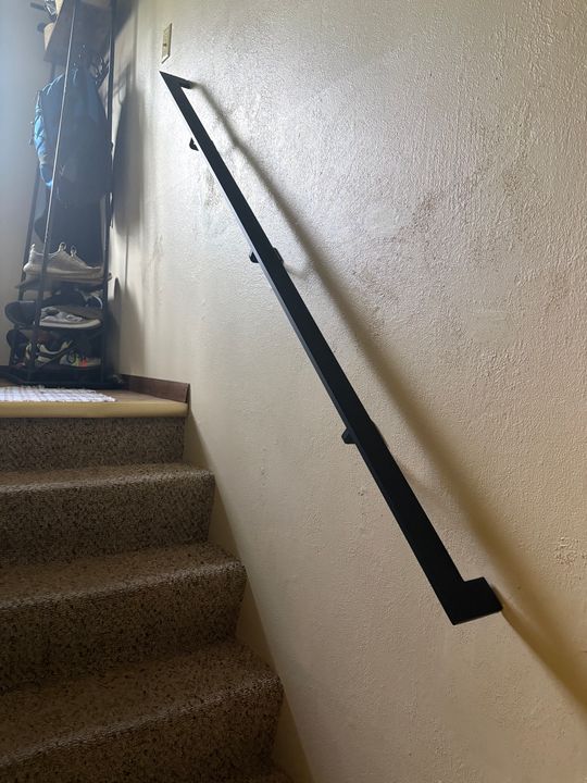 A black metal handrail mounted on a white wall alongside carpeted stairs