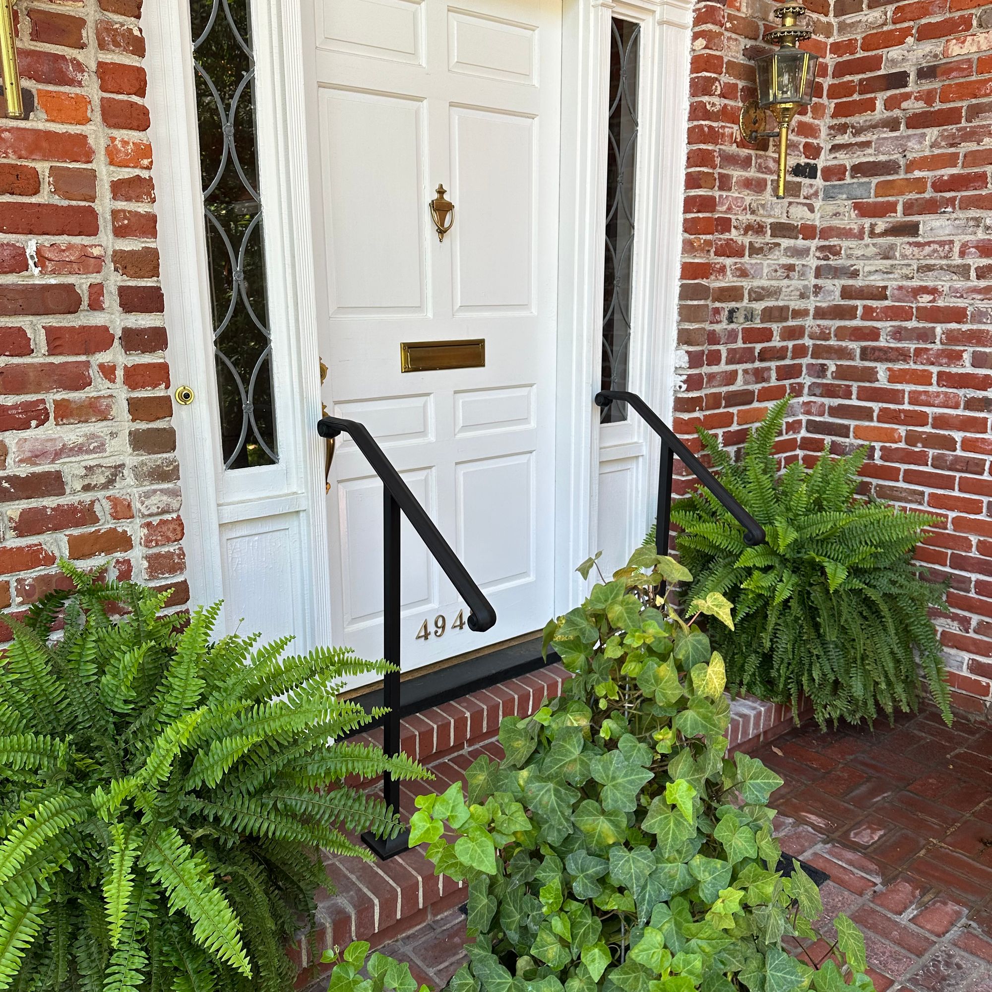 Front entrance of a brick home with white door, decorative sidelights, arched transom window, black handrails, and lush green plants including ferns flanking the steps