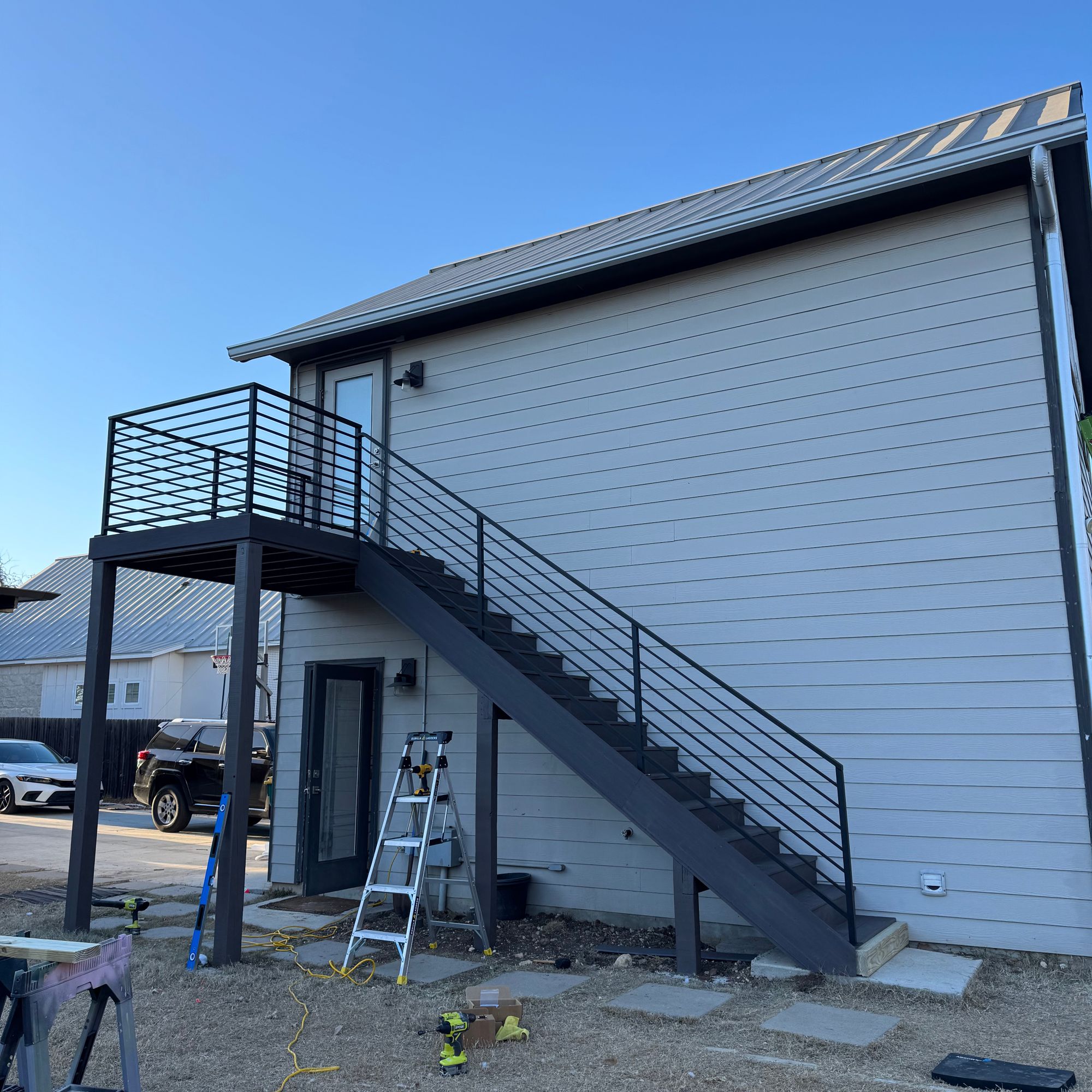 Modern two-story building with gray siding, external metal staircase with horizontal railings leading to upper level balcony, construction equipment and ladder visible on ground