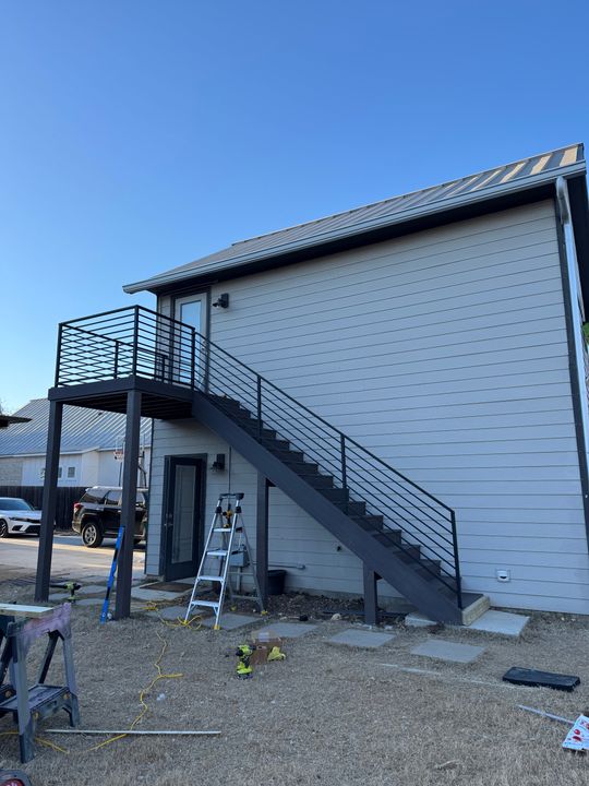 Modern two-story building with gray siding, external metal staircase with horizontal railings leading to upper level balcony, construction equipment and ladder visible on ground