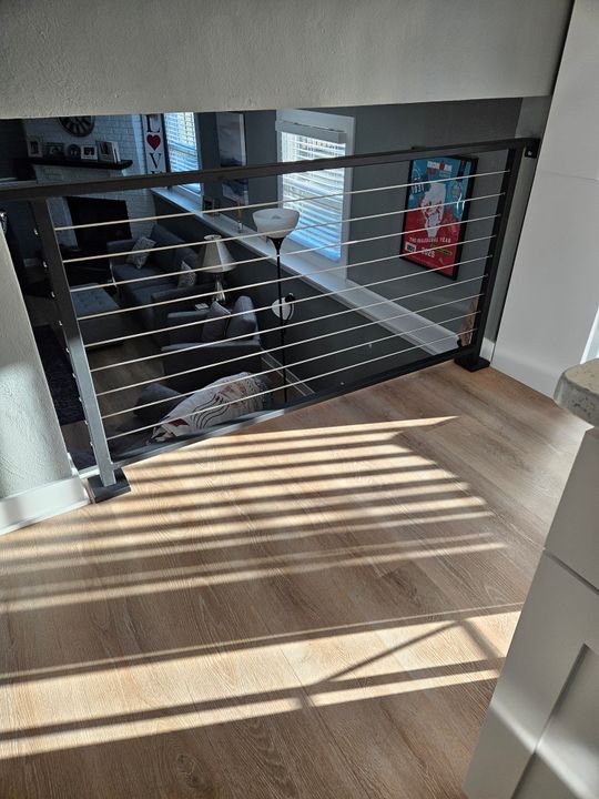 Interior view of a modern home showing a staircase landing with horizontal metal railings, wooden flooring with sunlight streaming through creating striped shadows, and a view down to the lower level living area with furniture and decor visible below.