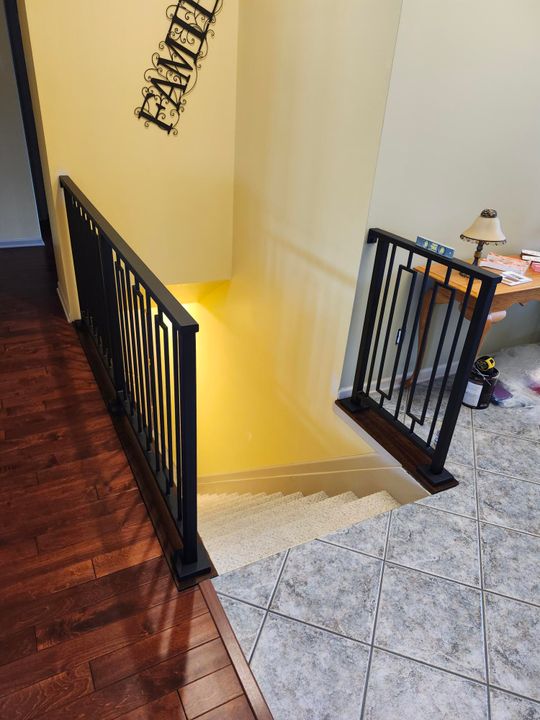 Interior staircase with black metal railings, hardwood flooring on upper level, and tile flooring on lower level with yellow accent lighting on the wall