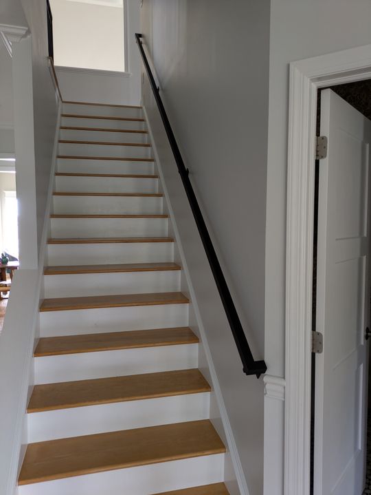 Interior staircase with white risers, natural wood treads, black handrail, and gray walls leading to upper floor