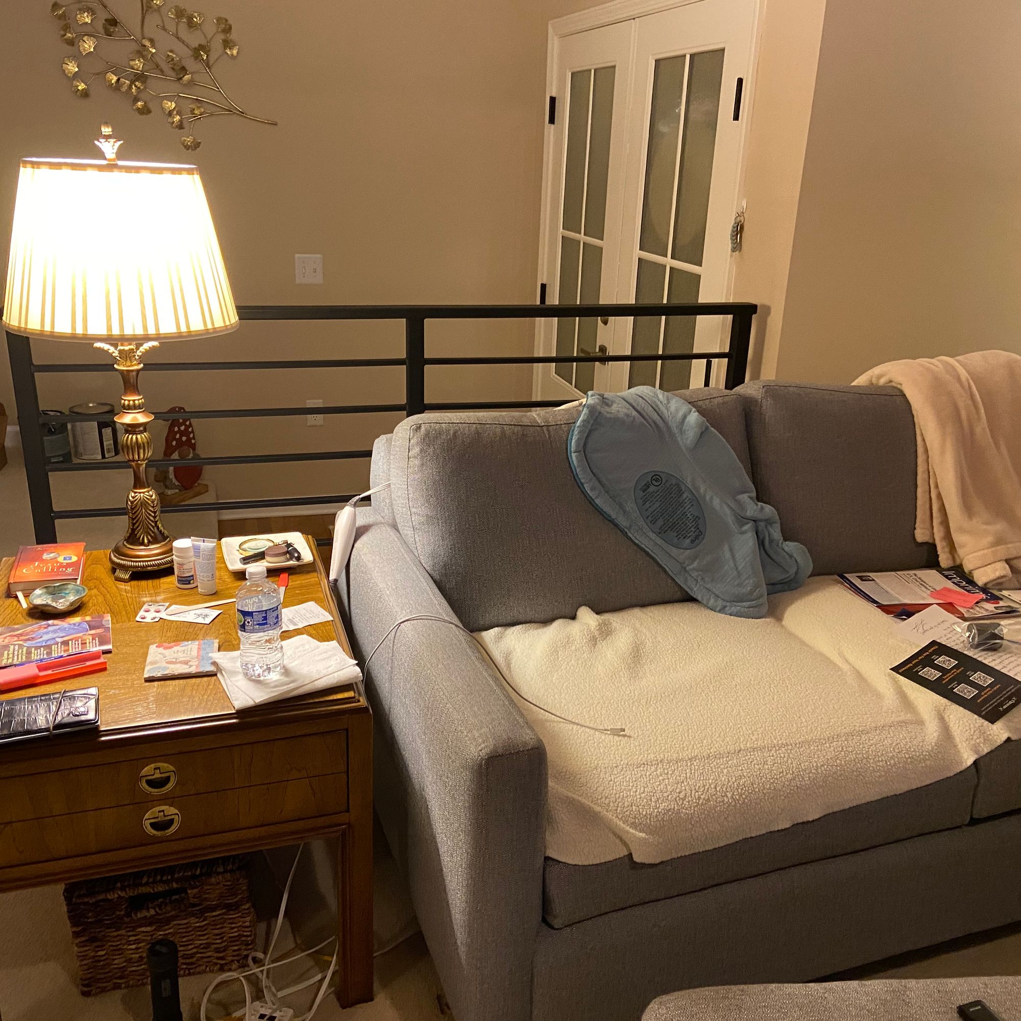 A cozy living room with a gray sectional sofa, wooden side table with lamp, magazines and books scattered on furniture, and warm lighting from a table lamp with pleated shade