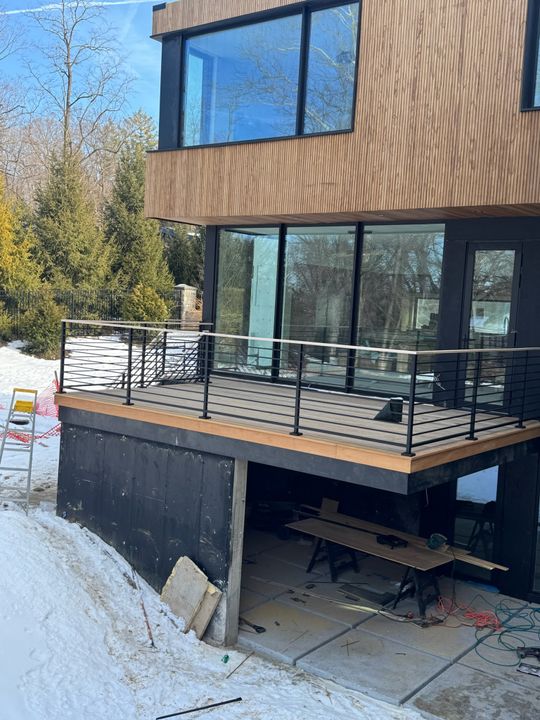 Modern two-story house with wood siding, large windows, and elevated deck with black railings under construction in winter