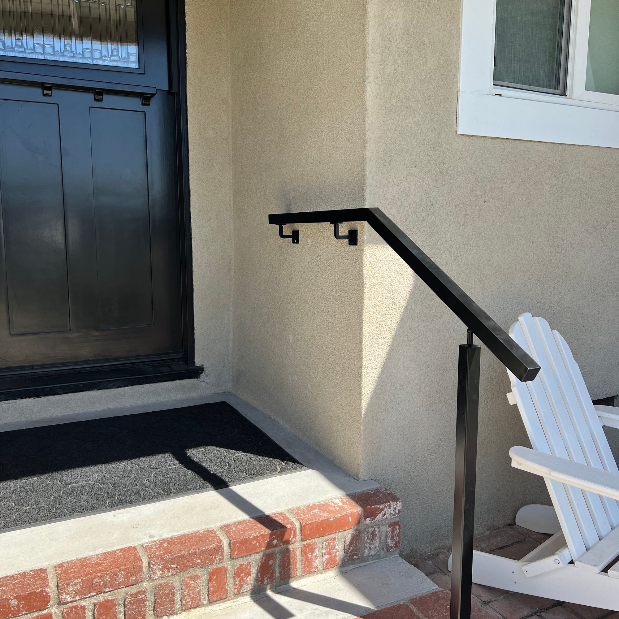 Front entrance of a house with brick steps, black metal handrail, blue door with decorative glass panel, and white Adirondack chair on brick patio