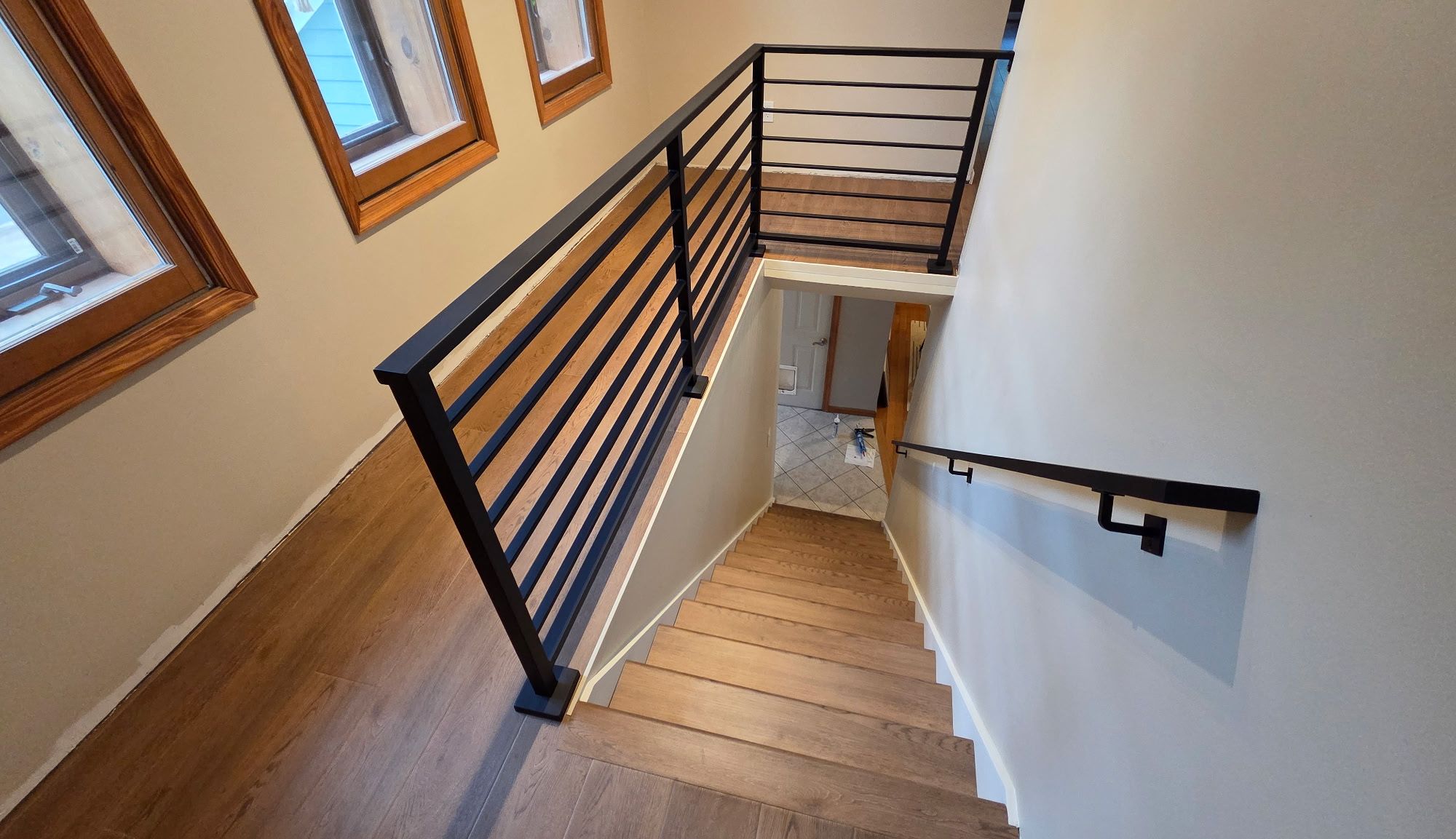 Modern wooden staircase with black metal horizontal railings leading downward, featuring wood-framed windows and white walls