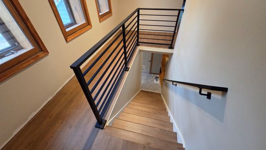 Modern wooden staircase with black metal horizontal railings leading downward, featuring wood-framed windows and white walls
