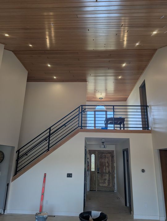 Modern interior with wooden plank ceiling, white walls, black metal staircase with horizontal railings leading to upper level, and front door visible in background