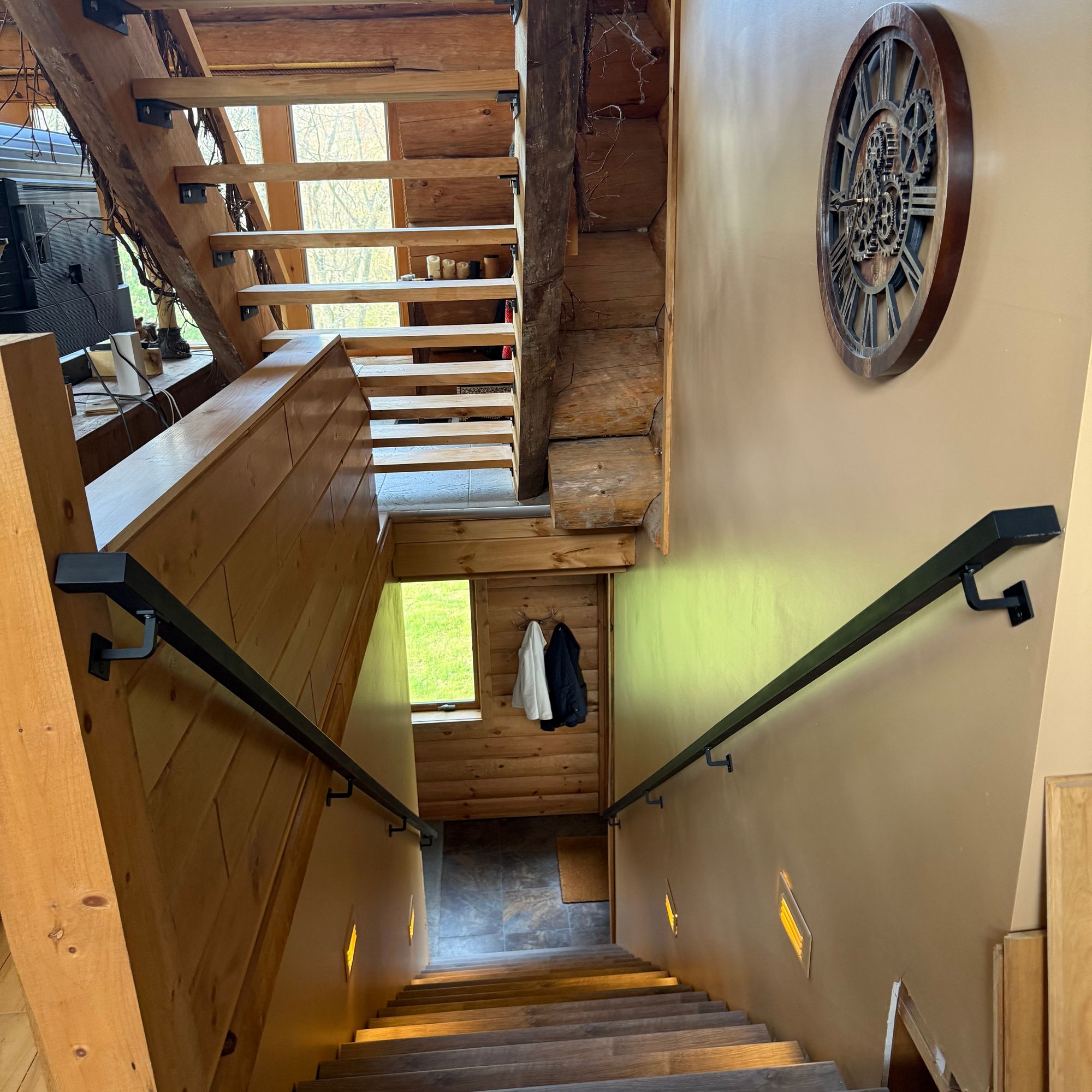 Interior view of a wooden staircase with black handrails leading upward, featuring exposed wooden beams, a decorative wall clock, and natural lighting from windows above