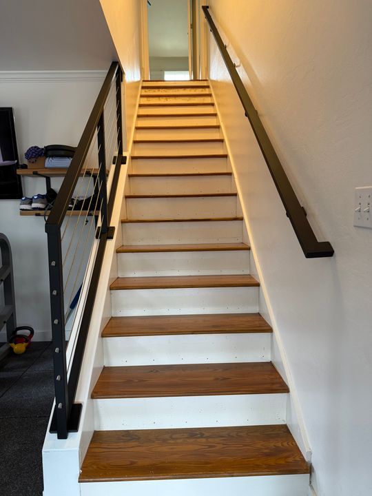 A modern staircase with wooden treads, white risers, and black metal cable railings leading to an upper floor in a contemporary home interior.