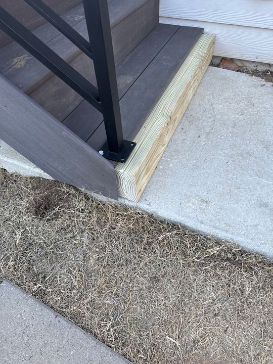 A wooden beam placed on concrete steps next to a black metal railing, with dried grass visible in the foreground