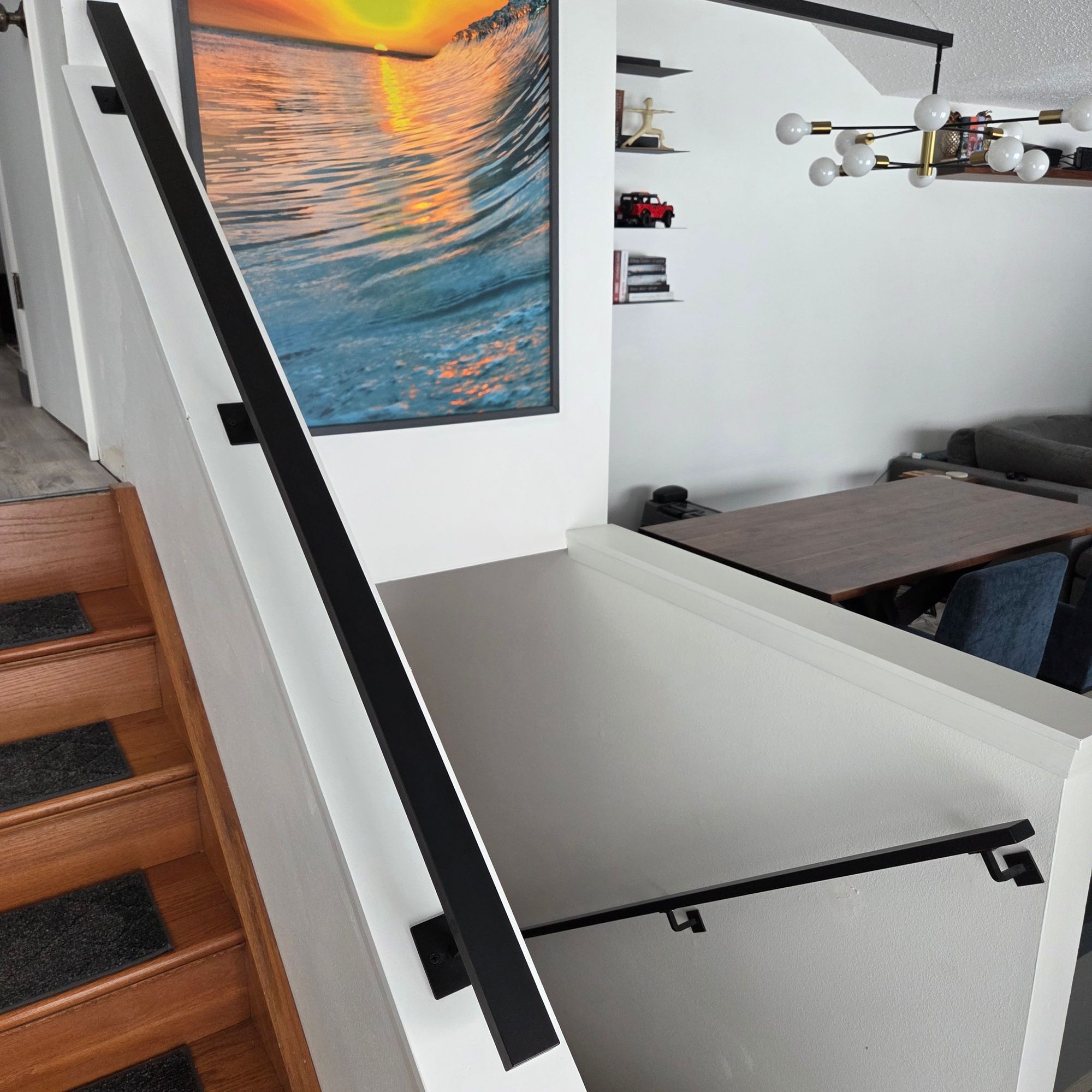 Modern staircase with white railings and black handrails, featuring a colorful ocean wave artwork on the wall and view of dining area below