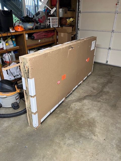 A large flat cardboard package or box standing upright in a garage with shelving, tools, and storage items visible in the background