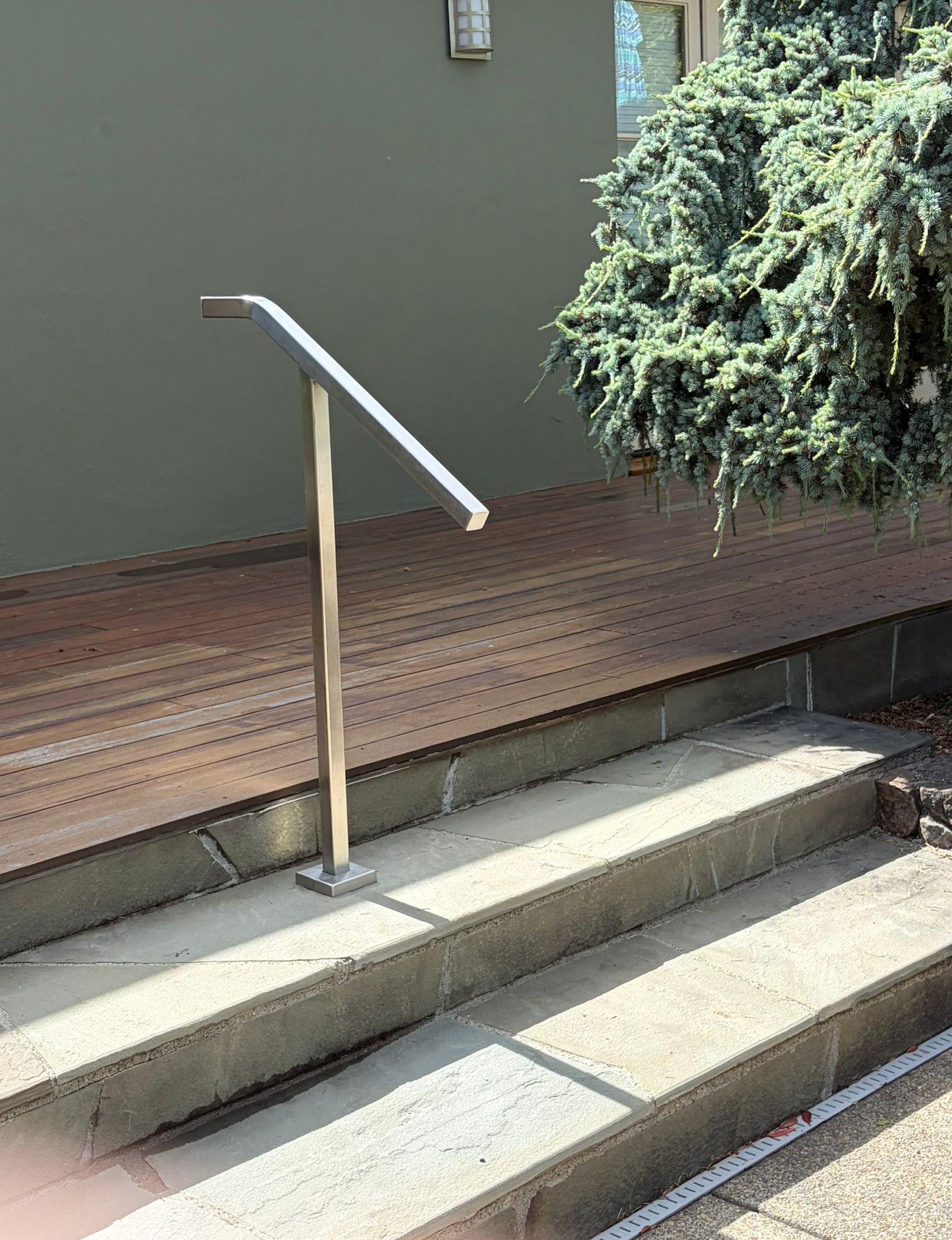 Modern metal handrail on concrete steps leading to wooden deck with evergreen shrub and gray building wall in background