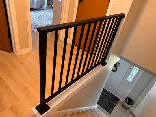 Interior staircase with dark wooden railing and vertical spindles overlooking an entryway with white door and dark floor mat