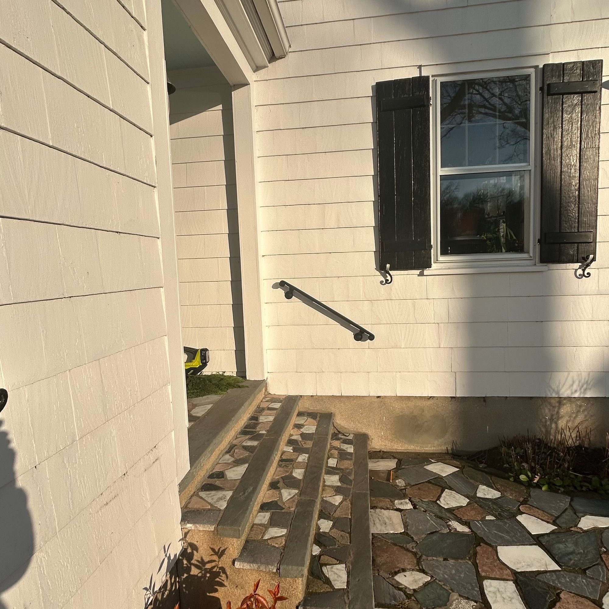 Exterior view of a white house with dark shutters, stone steps leading to an entrance, and a decorative stone patio with mixed colored tiles
