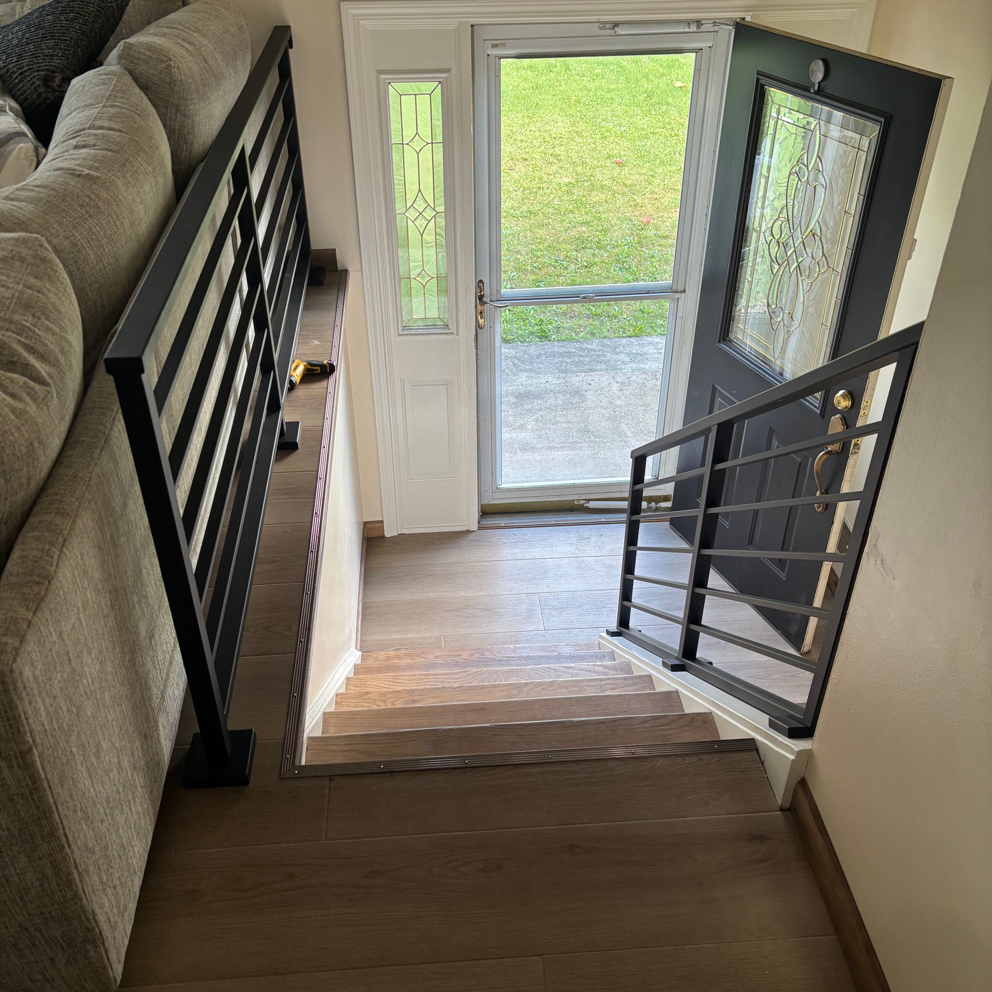 Interior view of a staircase with black metal railings leading down to an entryway with a white front door featuring decorative glass panels and sidelights, showing a green lawn outside