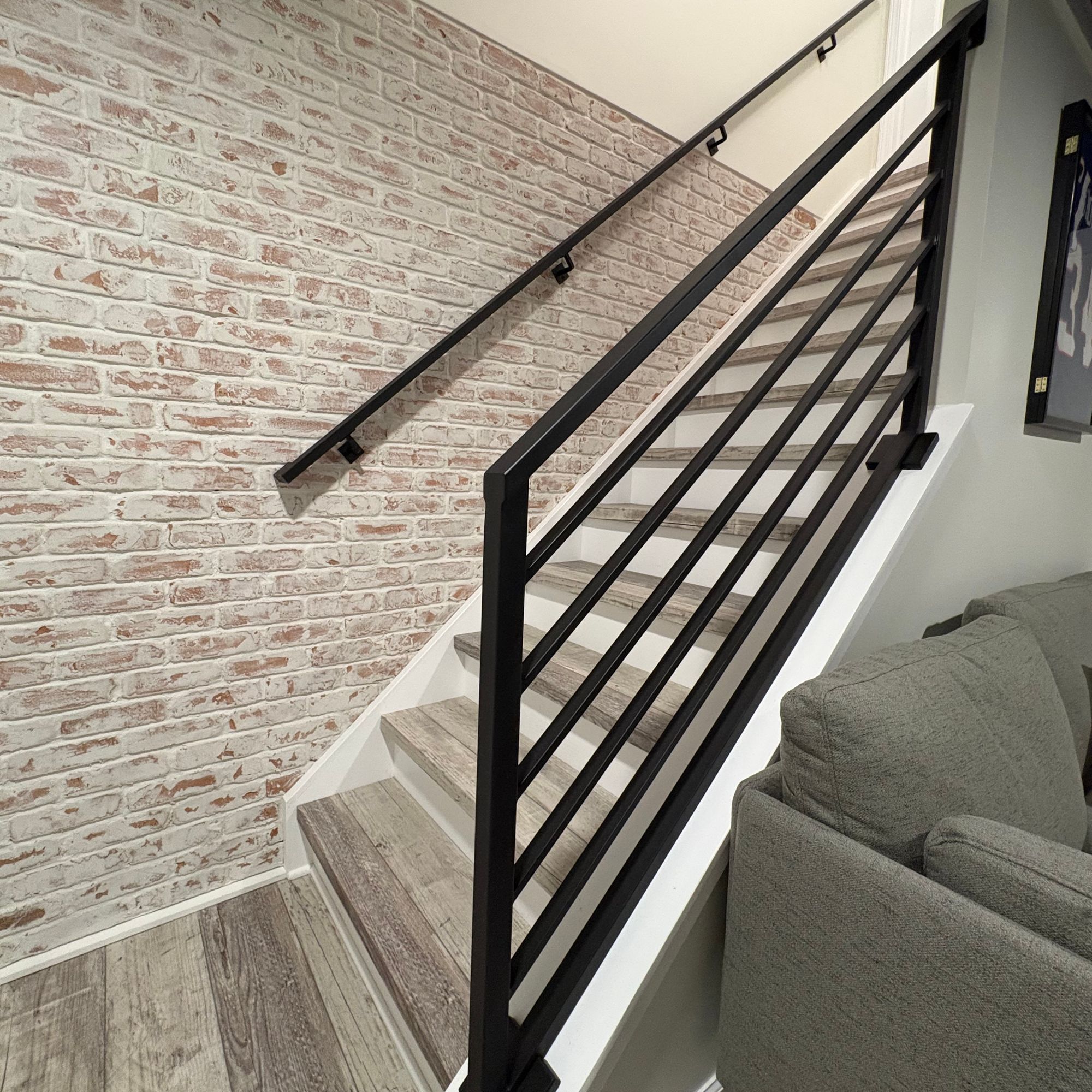 Modern staircase with black metal railing and white steps leading upward, featuring exposed brick wall and gray couch in foreground