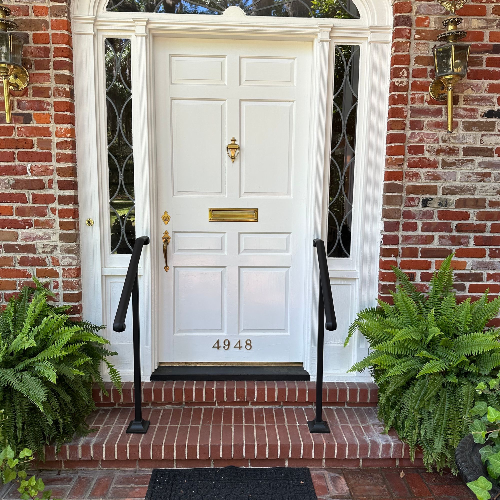 White front door with brass hardware and glass sidelights, surrounded by red brick archway, flanked by ferns and featuring black handrails leading to brick steps