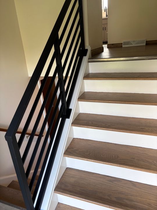 Modern staircase with wooden treads, white risers, and black metal geometric railing leading to upper floor