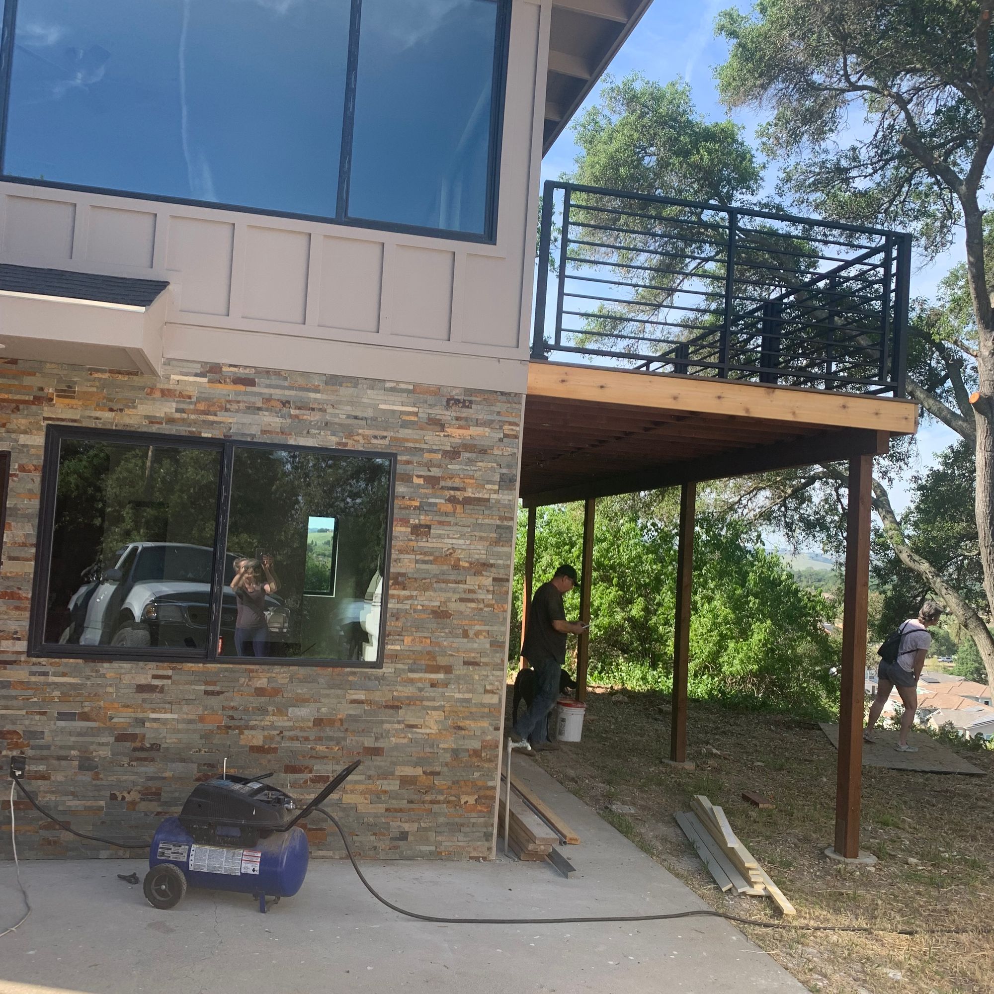 Modern two-story house under construction with stone and wood siding, featuring a wooden deck with metal railings, large windows, and construction equipment on a concrete patio surrounded by trees