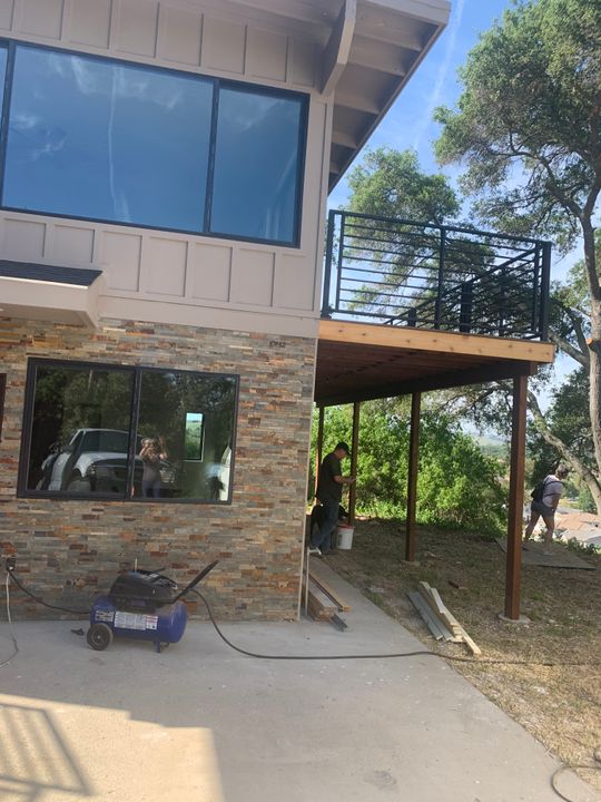Modern two-story house under construction with stone and wood siding, featuring a wooden deck with metal railings, large windows, and construction equipment on a concrete patio surrounded by trees