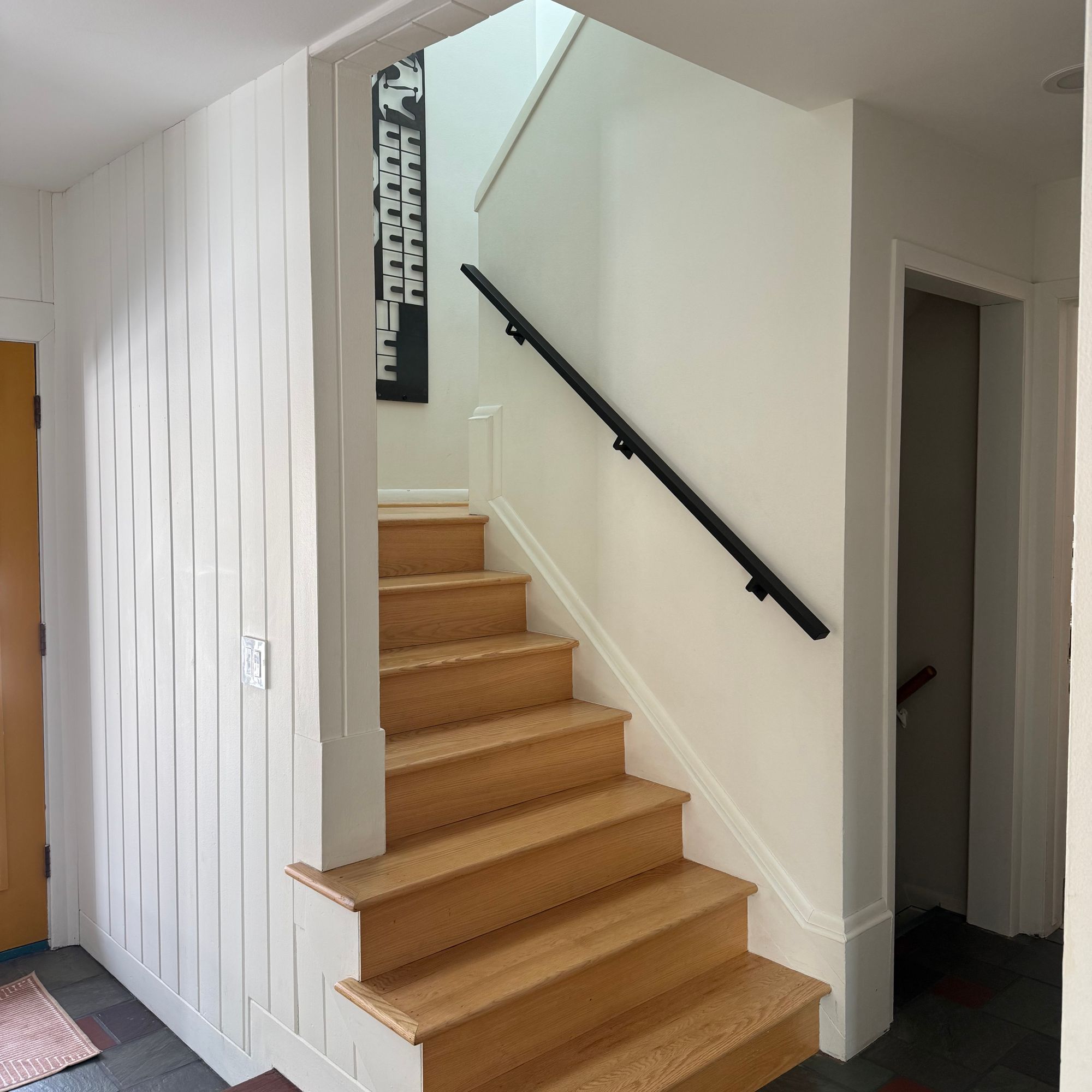 Interior staircase with wooden steps, black handrail, white walls, and multicolored tile flooring in a modern home entryway