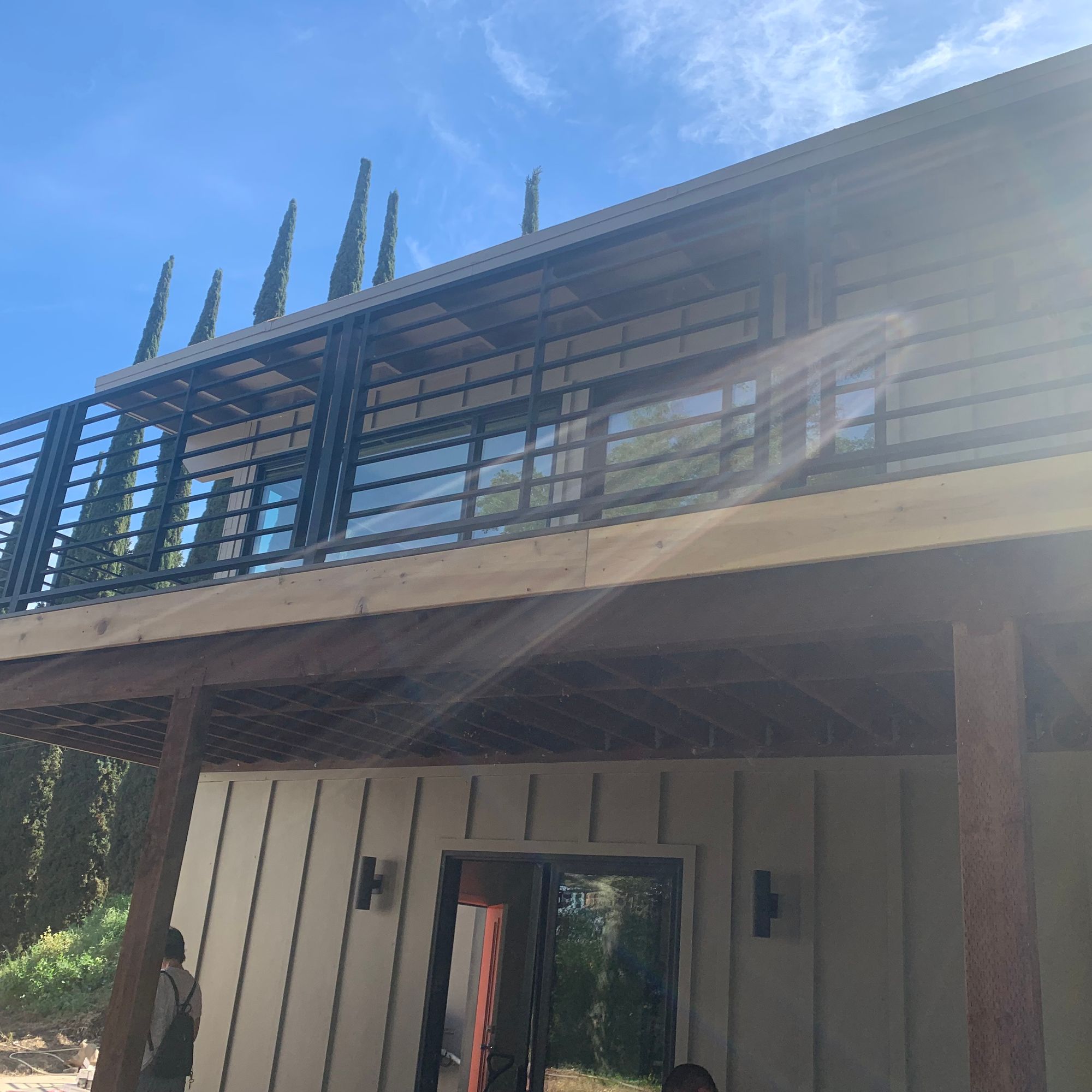 Modern two-story house under construction with metal railings on upper deck, wooden siding, and large windows against blue sky