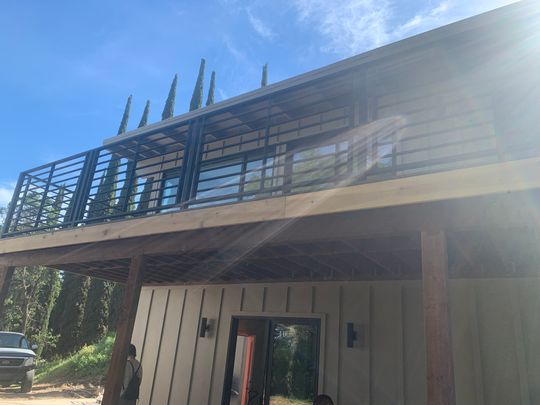 Modern two-story house under construction with metal railings on upper deck, wooden siding, and large windows against blue sky