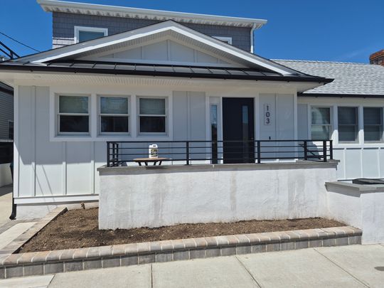 Modern single-story house with white and gray exterior, solar panels on roof, front porch with metal railing, and landscaped front yard with brick edging