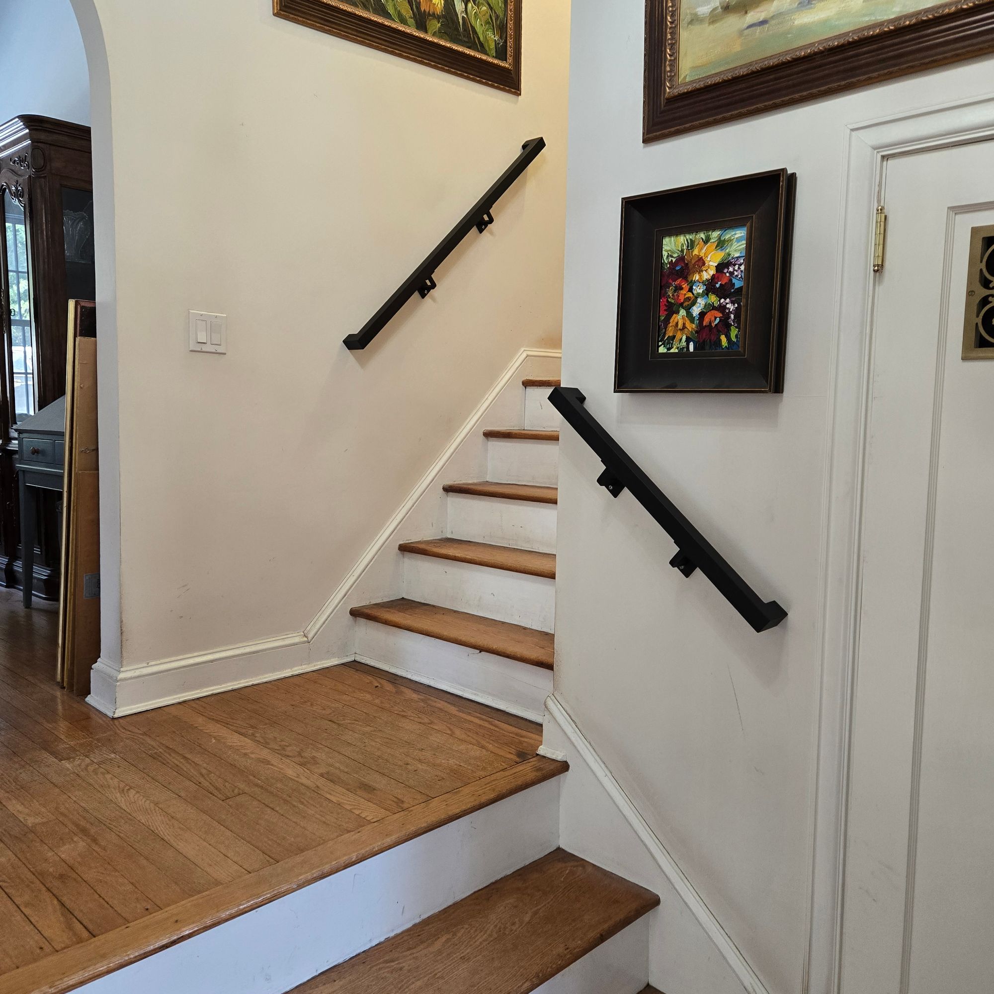 Interior staircase with wooden steps, white risers, and black handrails, decorated with framed artwork on cream-colored walls