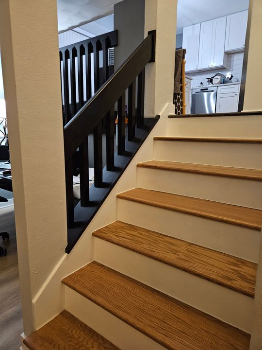 Interior staircase with wooden treads and black railings leading to upper level, with white kitchen cabinets and stainless steel appliances visible in background