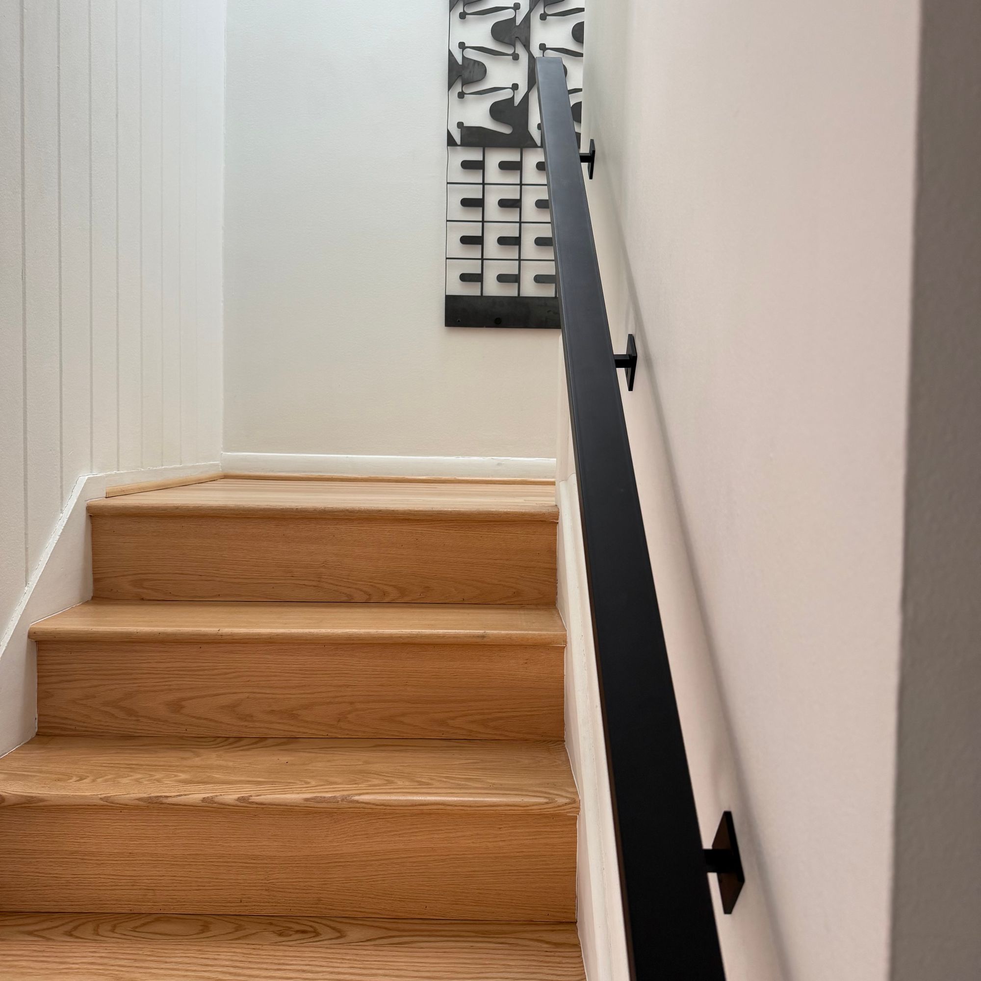 Wooden staircase with black metal handrail leading upward, white walls with decorative artwork visible at the top