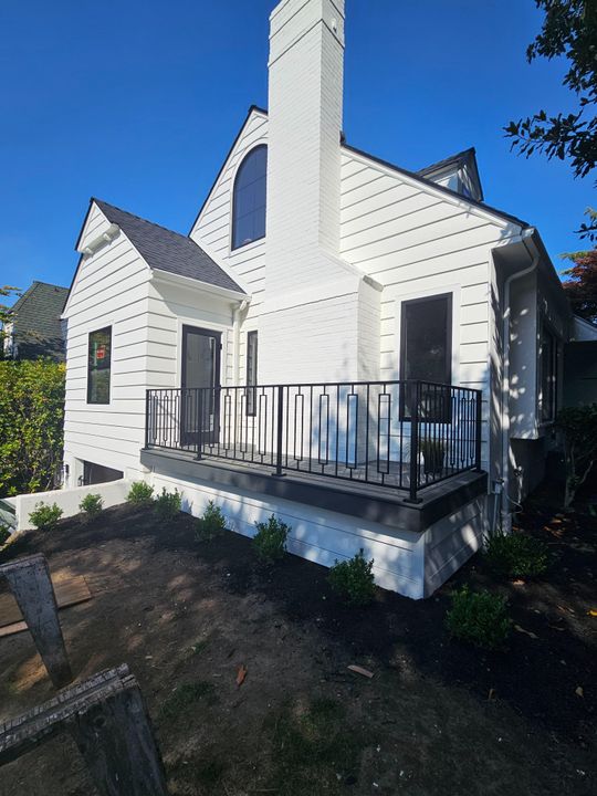 A white two-story house with dark roof, featuring a front porch with black metal railings, white chimney, and landscaping in the front yard under a clear blue sky.